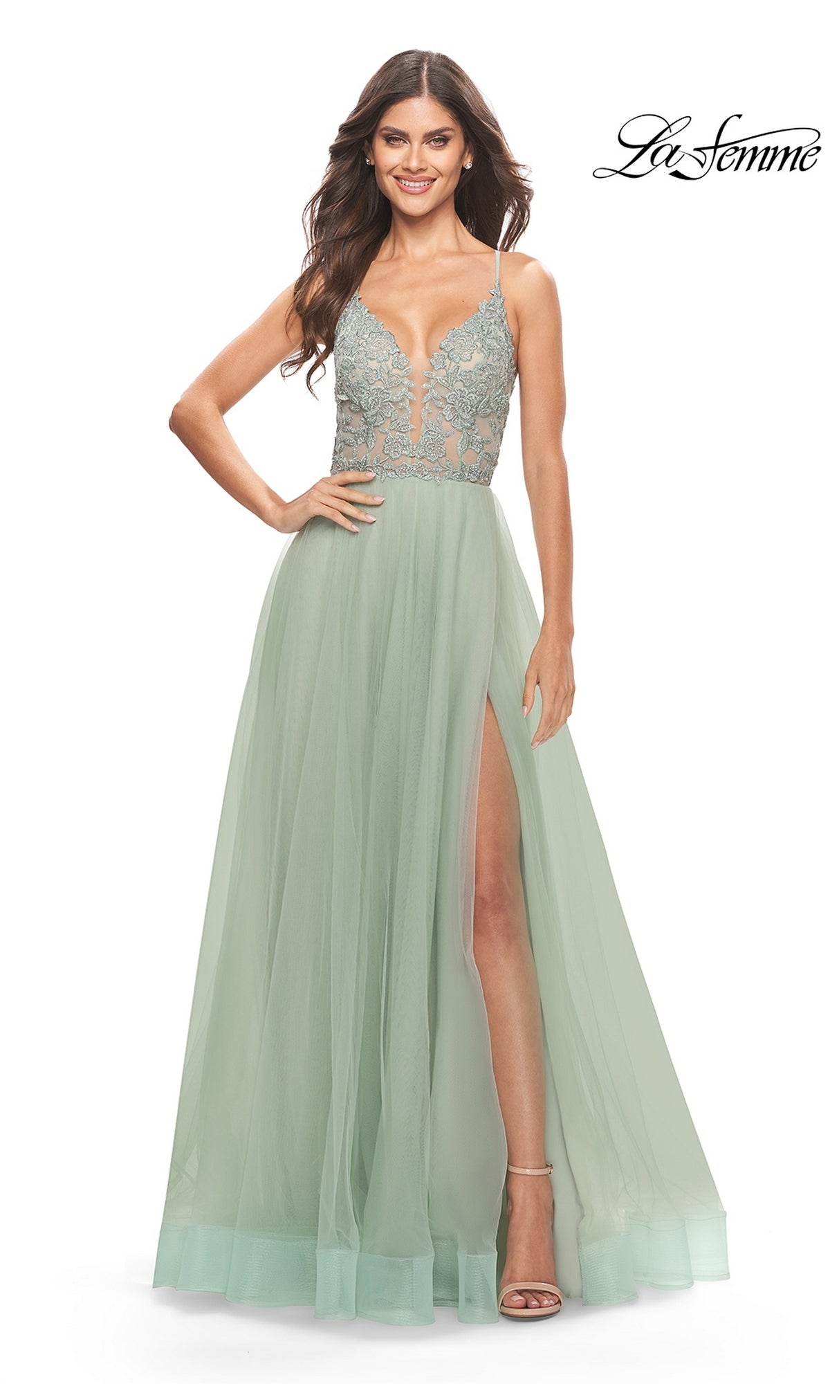 A woman wearing the Lace-Bodice Long A-Line Prom Dress La Femme 31542, featuring a light green hue, embroidered lace bodice, sheer skirt with high slit, and spaghetti straps, stands smiling against a white background.